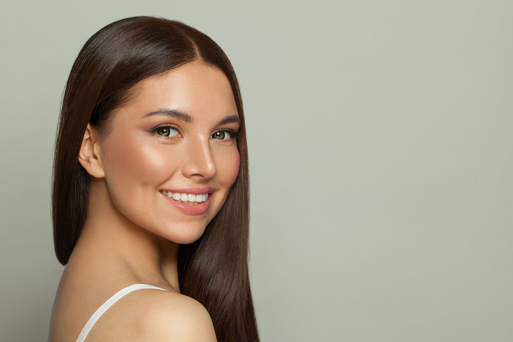 woman with long brown hair and smooth skin smiling slightly while looking over her shoulder against a plain background