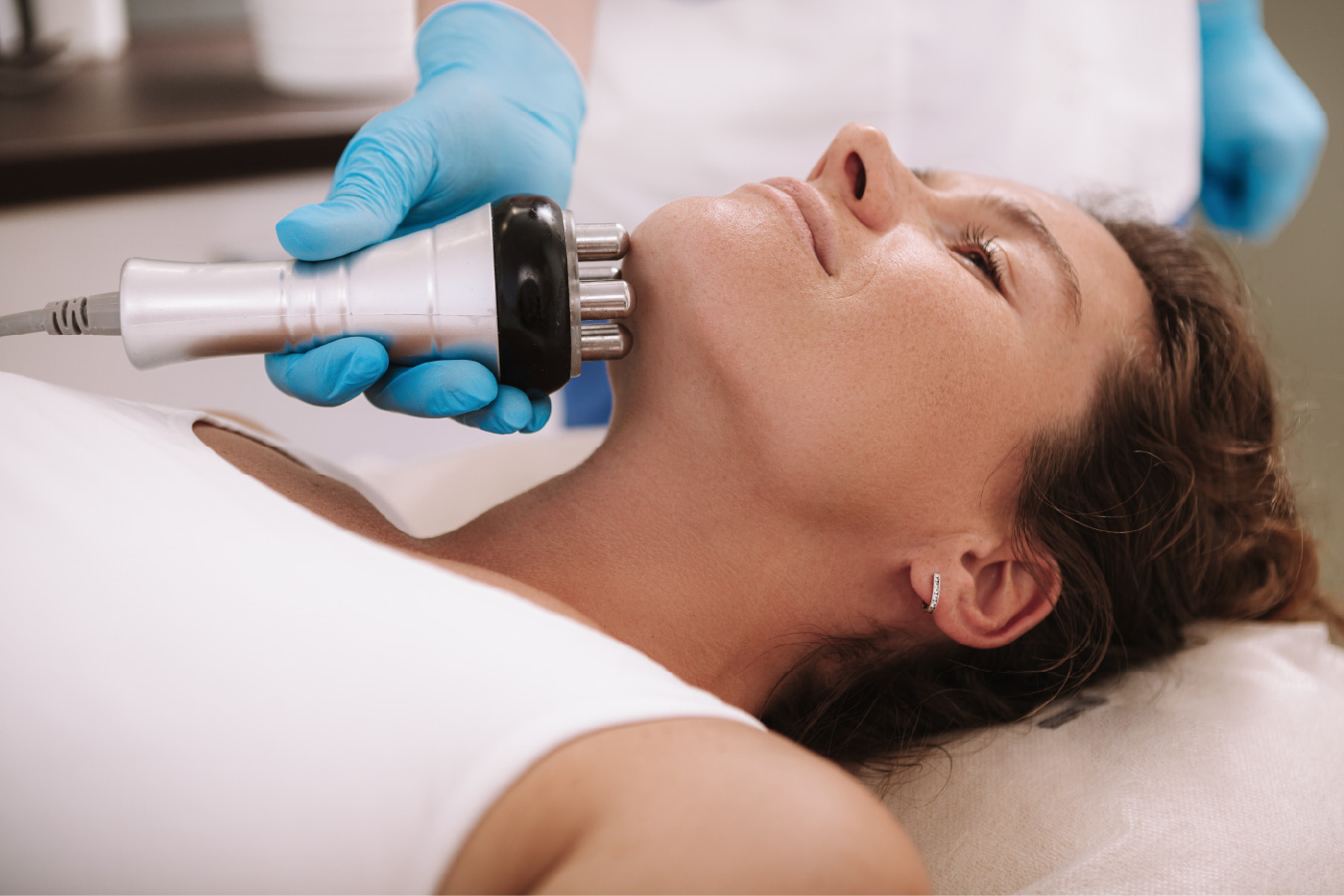 Gloved hand holding a multi-prong radiofrequency device against a woman’s neck as she lies on a treatment bed.