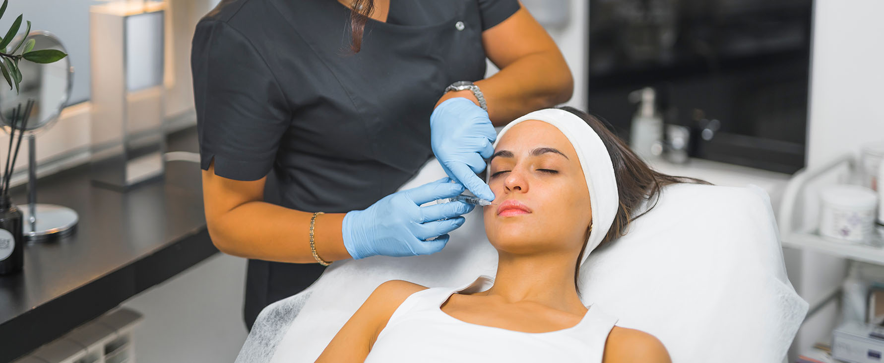 practitioner wearing blue gloves holding a syringe near a woman’s lips as she lies down