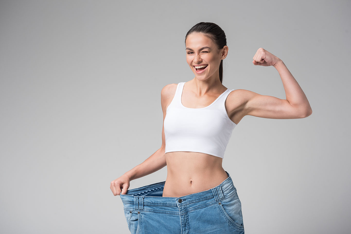 Woman holding the waistband of loose jeans with one hand while flexing her arm and winking.
