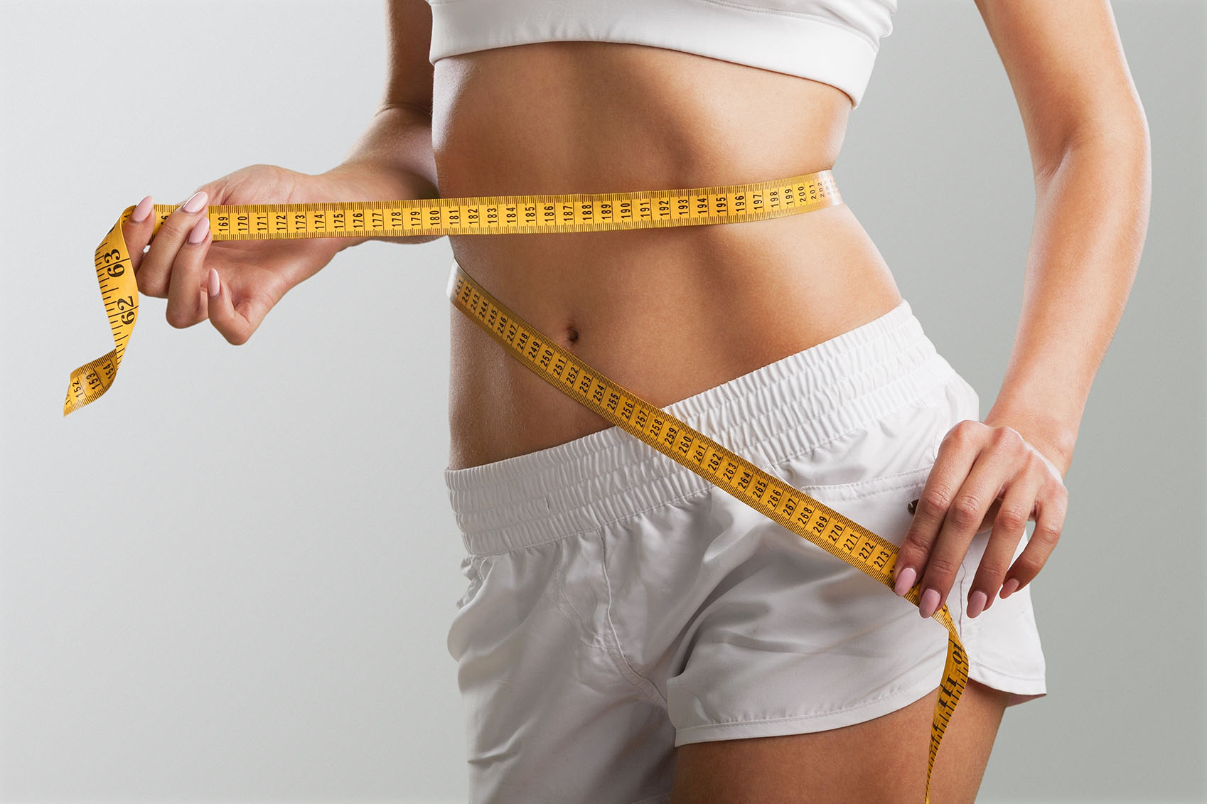 Close-up of a woman holding a yellow measuring tape around her waist while wearing white shorts.