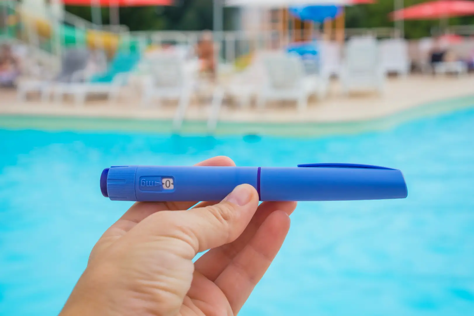Hand holding a blue injection pen in front of a swimming pool and lounge chairs.