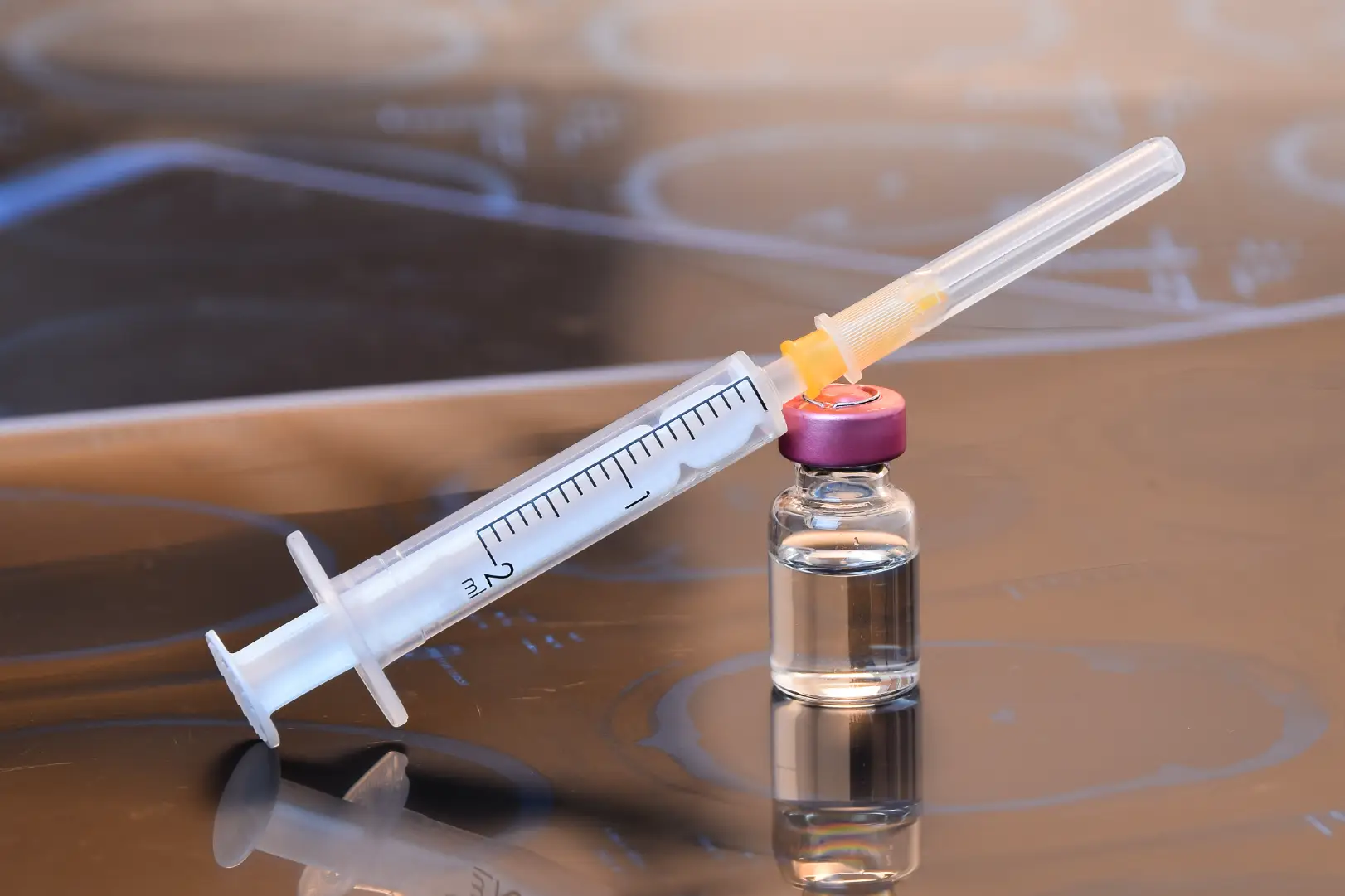 Syringe resting against a small vial filled with clear liquid on a reflective surface.