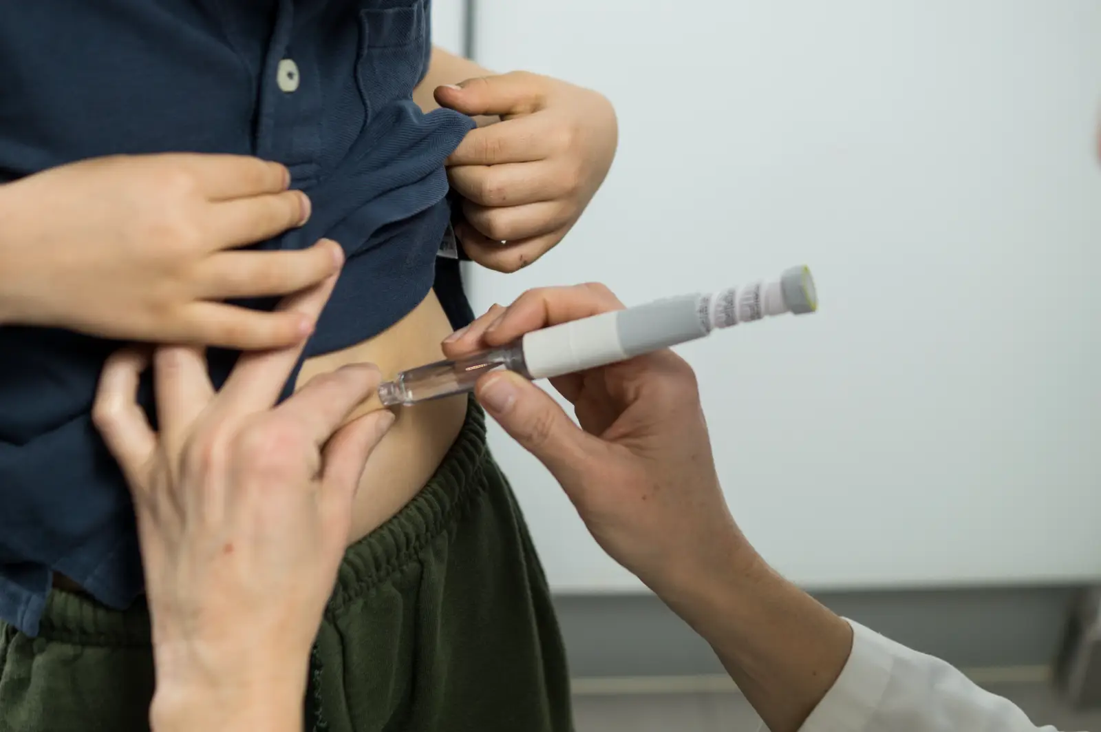 Hands hold an injection device against a person's abdomen while another hand lifts a shirt.