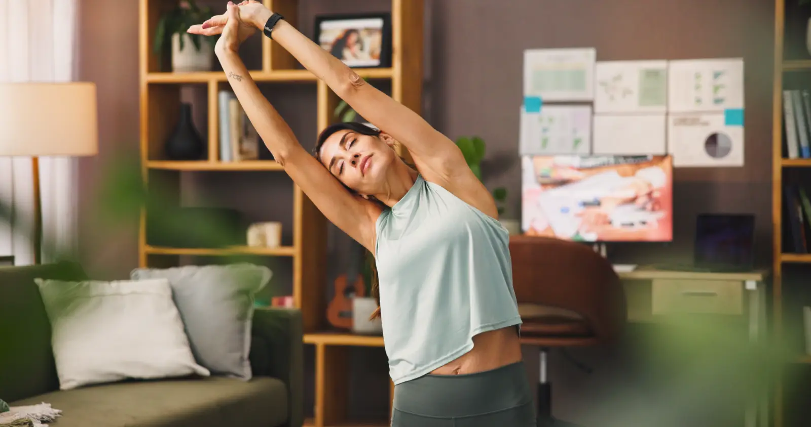 Person stretching with arms overhead in a room with shelves, a sofa, and a desk.